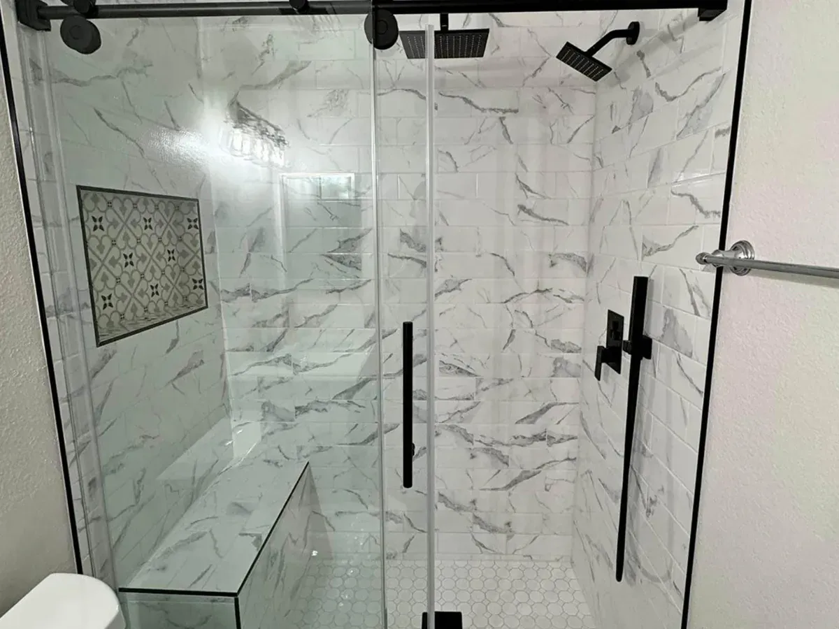 Frameless glass shower door with a black handle over marble look tile and a built-in bench in a Pasadena, MD bathroom